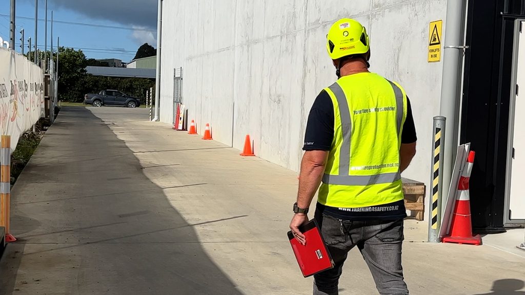 A man wearing high-vis and a hard hat is walking along the side of a concrete building holding a clipboard